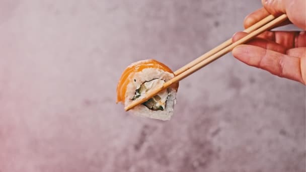 寿司の棒を使って寿司ロールを1本取り 閉じます お箸を持った人の手は サーモンとフィラデルフィアチーズがカラフルな背景で新鮮な日本の寿司を示しています 魚介類 — ストック動画