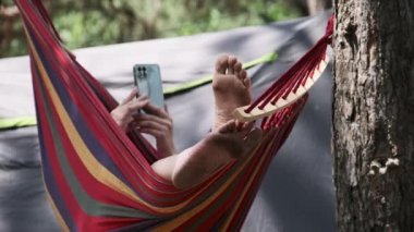 Bir kadın turist çadırın yanındaki hamakta uzanıyor ve akıllı telefon kullanıyor. Dişi ayaklarının görüntüsü. Hamakta sallanan ve yaz kampı yakınlarındaki bir ormanda sosyal medyada sallanan biri. Tatil günü. 4K