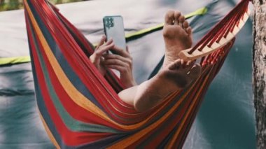 Bir kadın turist çadırın yanındaki hamakta uzanıyor ve akıllı telefon kullanıyor. Dişi ayaklarının görüntüsü. Hamakta sallanan ve yaz kampı yakınlarındaki bir ormanda sosyal medyada sallanan biri. Tatil günü. 4K