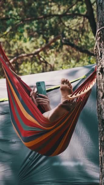 サマーキャンプ場でハンモックに乗りながらスマートフォンを使った垂直な女性 女性旅行者はテントの近くの松林で携帯電話を使用しています ソーシャルネットワークのチェック リラックス ライフスタイル — ストック動画