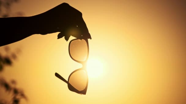 女人手拿太阳镜挡住日落的天空 带着深色眼镜的女性手臂在天空中的轮廓 太阳光穿过太阳镜 享受户外运动 自然和生活方式 — 图库视频影像