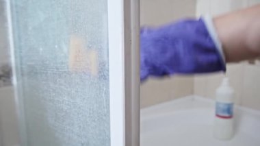 Banyo temizliği ağır çekimde. Mavi lastik eldivenli bir kadın turuncu bir süngerle duş kabininin kirli yüzeyini temizliyor. Dişi eller banyoyu siliyor. Pürüzsüz kamera hareketi. Ev işi temizliği.