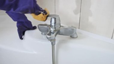 Mavi lastik eldivenli bir kadın kirli banyoyu ağır çekimde turuncu bir süngerle temizliyor. Banyo temizleyen kadın ellerinin yakın çekimi. Krom suyu musluğunu yıkıyor. Ev işi temizliği.
