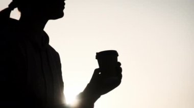 Genç bir kadının silueti, güzel bir günbatımı gökyüzünde yavaş çekimde sıcak kahve içer. Güneş ışınları batan bir kız, elinde kahveyle parktaki bir korkuluğa oturur ve hayatın tadını çıkarır. Sağlıklı yaşam tarzı