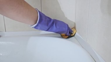 Kirli tuvaleti turuncu süngerle yavaş çekimde temizleyen bir kadın. Mavi lastik eldivenli kadın elleri banyoyu siliyor. Küvet yüzeyini deterjanlarla temizliyorum. Ev işi, temizlik konsepti