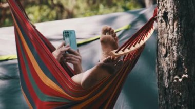 Yaz kampında hamakta sallanırken akıllı telefon kullanan bir kadın. Kadın gezgin çadırın yakınındaki bir çam ormanında cep telefonu kullanıyor. Sosyal ağları kontrol etmek, çevrimiçi kalmak, rahatlamak, yaşam tarzı