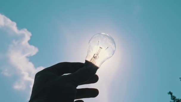 男性の手のPovシルエットは 明るい太陽と青い空に対して電球を保持しています 古い白熱した電球と日光 太陽光線は電球を通して光る 再生可能エネルギー — ストック動画