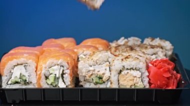 Yemek çubukları suşi kutusuna bir suşi koydu, yakın plan. Plastik bir kutuda yuvarlanıyor. Çeşitli lezzetli suşi setleri bir teslimat konteynerinde. Konsept Asya yemekleri, sağlıklı yiyecekler, paket yemek.