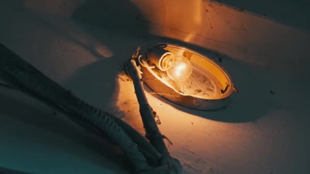 一盏白炽灯在水泥墙上的一盏破灯里发出光芒 房子里有危险电线的建筑物照明 楼梯井中有蜘蛛网的旧钨球 — 图库视频影像