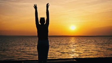 Genç ve rahat bir kadının silueti yavaşça günbatımının tadını çıkarırken ellerini kaldırıyor. Genç bir kızın batan güneşe bakışının arkası. Yaşam tarzı, özgürlük, mutluluk, akıl sağlığı kavramı