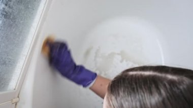 Kirli tuvaleti turuncu süngerle yavaş çekimde temizleyen bir kadın. Mavi lastik eldivenli kadın elleri banyoyu siliyor. Küvet yüzeyini deterjanlarla temizliyorum. Ev işi, temizlik konsepti