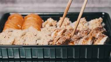 Bambu çubukları suşi kutusundan suşi alır, yakın plan. Plastik bir kutuda yuvarlanıyor. Çeşitli lezzetli Japon suşilerinden oluşan hazır yemek takımı bir teslimat konteynerinde yuvarlanıyor. Yemek teslimatı kavramı.