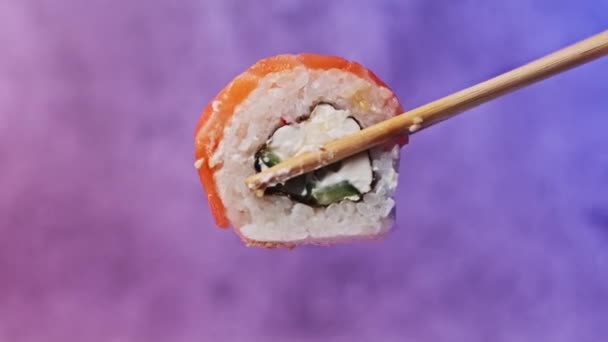 寿司の棒を使って寿司ロールを1本取り 閉じます お箸を持った人の手は サーモンとフィラデルフィアチーズがカラフルな背景で新鮮な日本の寿司を示しています 魚介類 — ストック動画