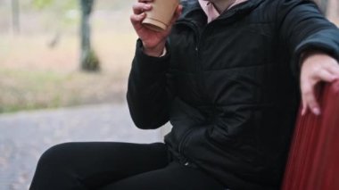 Genç bir kadın sonbahar parkındaki bankta kağıt bardaktan kahve içiyor. Kadın eli bir fincan aromatik kahve tutuyor. Ağır çekimde hayattan zevk alan bir kadın. Sonbahar mevsimi.