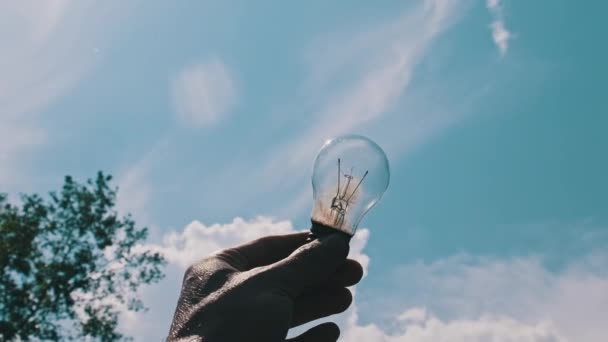 Pov男性の手は青空の明るい太陽に対して白熱電球を保持しています 腕のシルエットは 太陽の光を照らす電気ランプを保持しています アイデアソーラーエネルギー エコエネルギー 省エネルギー — ストック動画
