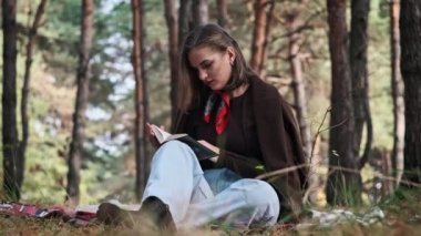 Genç bir kadın çam ormanında bir battaniyenin üzerinde otururken kitap okuyor. Makyajlı ve şık giyimli güzel kız dışarıda okumayı seviyor. Doğada dinlenmek. Sayfayı çevir. Ağır çekim. 4K.