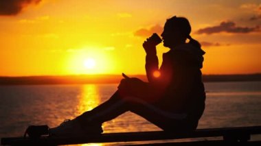 Genç kadın silueti, portakal rengi günbatımında sıcak kahve içer. Yavaş çekimde suyun yanında. Güneş ışınları batan bir kız elinde kahveyle korkuluğa oturur ve hayatın tadını çıkarır. Sağlıklı yaşam tarzı
