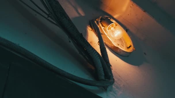 一个古老的白炽灯泡在楼梯井混凝土墙上的一盏破碎的灯上发出光芒 灯丝闪烁着黄色的光芒 隐隐约约地照在墙上挂着蜘蛛网的旧电盒里 — 图库视频影像