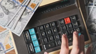Üst manzara, dolar banknotlarıyla çevrili eski model bir hesap makinesine güveniyor. Yakın plan. Eski bir hesaplama makinesinde erkek eli klavye tuşlarına basıyor. Finansal hesaplar, para birimi, tasarruf, 4K faturaları