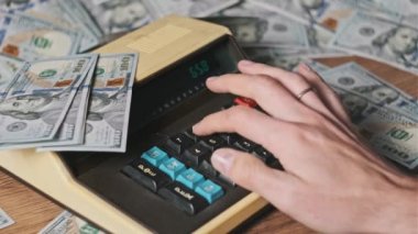 Dolarlarla çevrili eski bir hesap makinesine güvenerek, yakın plan. Eski bir hesaplama makinesinde erkek eli klavye tuşlarına basıyor. Finansal hesaplar, para birimi, tasarruflar, mali faturalar 4K