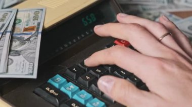 Dolarlarla çevrili eski bir hesap makinesine güveniyoruz. Yakın plan. Tahta masadaki eski hesaplama makinesinin klavye tuşlarına erkek eli basar. Konut finansmanı, para birimi, tasarruf, mali faturalar
