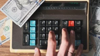 Üst görünüm, dolarlarla çevrili eski bir hesap makinesine güveniyor. Yakın plan. Tahta masadaki eski hesaplama makinesinin klavye tuşlarına erkek eli basar. Para, para, tasarruf, faturalar. 4K