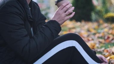 Genç bir kadın, sonbahar parkındaki çimlerin üzerinde düşmüş sarı yaprakların arasında oturup kahve içiyor. Kadın eli kağıt bardakta bir fincan aromatik kahve tutuyor. Hayatın tadını çıkarıyor, sakin sakin. Yavaş çekim.