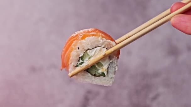 寿司の棒を使って寿司ロールを1本持ってきて握ると 背景がぼやけてしまい 閉じます 箸で人間の手はサーモンとフィラデルフィアチーズと新鮮な日本の寿司を示しています 魚介類 — ストック動画