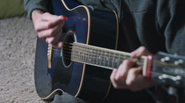 Genç adam evde akustik gitar çalar. Oturma odasında kanepede oturan müzisyen gitarı. Şarkı çalmayı öğrenmek, telleri tıngırdatmak, akorlar çalmak. Rahatlatıcı, yaşam tarzı konsepti, akıl sağlığı.