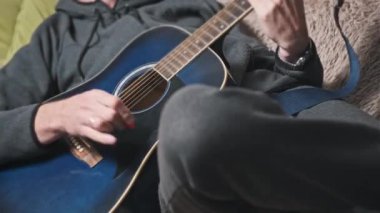 Genç adam evde akustik gitar çalar. Oturma odasında kanepede oturan müzisyen gitarı. Şarkı çalmayı öğrenmek, telleri tıngırdatmak, akorlar çalmak. Rahatlatıcı, yaşam tarzı konsepti, akıl sağlığı.