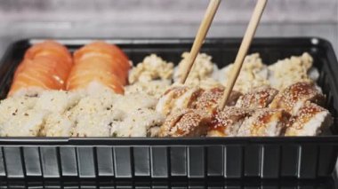 Sopalar suşi kutusundan suşi alır, yakın plan. Plastik bir kutuda yuvarlanıyor. Çeşitli lezzetli Japon suşilerinden oluşan hazır yemek takımı bir teslimat konteynerinde yuvarlanıyor. Yemek teslimatı kavramı. Paket yemek.