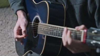 Genç adam evde akustik gitar çalar. Oturma odasında kanepede oturan müzisyen gitarı. Şarkı çalmayı öğrenmek, telleri tıngırdatmak, akorlar çalmak. Rahatlatıcı, yaşam tarzı konsepti, akıl sağlığı.