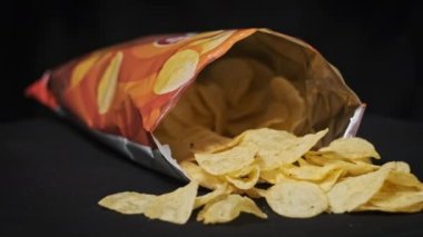 Açık bir patates cipsi paketi siyah arka planda yakın plan çekimde döner. Masaya birkaç cips dökülmüş bir paket açık cips. Fast food, abur cubur, sağlıksız yiyecekler. 4K