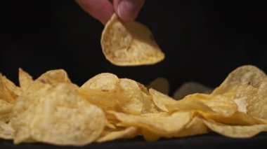 Erkek eli, patates cipsini siyah arka planda, Yavaş Hareket 'te yakın plan çeker. Erkek parmaklar masadaki bir yığın kızarmış cipsten cips alır. Fast food, abur cubur, sağlıksız yemek yemek.
