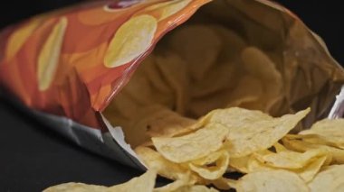 Açık bir fiş paketi siyah arka planda yakın plan çekimde döner. Açık patates cipsi torbası ve masaya birkaç cips dökülmüş. Fast food, abur cubur, sağlıksız yiyecekler