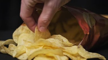 Erkek eli, siyah arka planda yakın plan patates cipsi alır. Erkek parmaklar, masada açık bir paketin yanında duran bir yığın cipsten atıştırmalık alır. Fast food, abur cubur, sağlıksız yemek yemek.