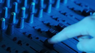 DJ el, konsol faderlarını neon ışıklı yakın planda karıştırmak için ses seviyesini ayarlar. Ses mühendisi ses karıştırıcıda düğmeleri aşağı yukarı hareket ettiriyor. Renkli arka planda çalışan adam müzik çalar