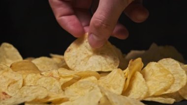 Erkek el, patates cipsini siyah arka planda yakın plan Yavaş Hareket 'e alır. Erkek parmakları, masadaki cips yığınından çerez alır. Fast food, abur cubur, sağlıksız yemek yemek.