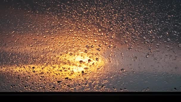 雨はゆっくりと窓のガラスから日没の空に向かって流れます オレンジ色と黄色の空が輝き ドロップレットを閉じる光を照らしています 雨水のパターンが落ちる スペースのコピー テキストの背景 — ストック動画