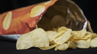 Açık bir patates cipsi paketi siyah arka planda yakın plan çekimde döner. Masaya birkaç cips dökülmüş bir paket açık cips. Fast food, abur cubur, sağlıksız yiyecekler. 4K