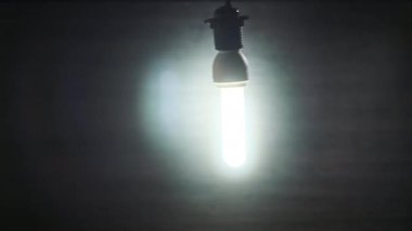 Spiral flüoresan lamba yanıp sönüyor, ışıkları yanıyor ve yakın plan bir elektrik telinde sallanıyor. Tüp ampulü koyu arkaplanda açılıp kapanıyor. Fikir, uzayı kopyalamak. Elektrik enerjisi tasarruf ampulü. 4K