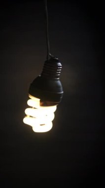 Dikey spiral floresan lamba yanıp sönüyor, ışıkları yanıyor ve yakın plan elektrik telinde sallanıyor. Tüp ampulü koyu arkaplanda açılıp kapanıyor. Fikir, uzayı kopyalamak. Elektrik enerjisi tasarruflu ampul 4K