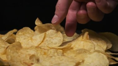 Erkek eli, patates cipsini siyah arka planda, Yavaş Hareket 'te yakın plan çeker. Erkek parmaklar masadaki bir yığın kızarmış cipsten cips alır. Fast food, abur cubur, sağlıksız yemek yemek.
