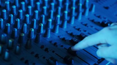 DJ el, konsol faderlarını neon ışıklı yakın planda karıştırmak için ses seviyesini ayarlar. Ses mühendisi ses karıştırıcıda düğmeleri aşağı yukarı hareket ettiriyor. Renkli arka planda çalışan adam müzik çalar