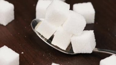 Kaşıktaki şeker küpleri masanın yakın çekiminde düzgün döner. Bir yığın tatlı beyaz şekerli metal kaşık. Kavramsal şeker bağımlılığı, sağlıksız beslenme, aşırı kilo, diyabet ve obezite.