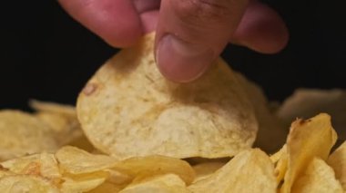 Erkek eli, patates cipsini siyah arka planda, Yavaş Hareket 'te yakın plan çeker. Erkek parmaklar masadaki bir yığın kızarmış cipsten cips alır. Fast food, abur cubur, sağlıksız yemek yemek.
