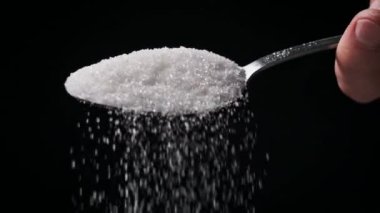 Siyah arka planda kaşıktan şeker düşüyor Ağır çekim, yakın plan. Bir çorba kaşığı granüle şeker ölç. Şeker bağımlılığı, sağlıksız beslenme, aşırı kilo, diyabet, obezite. Boşluğu kopyala