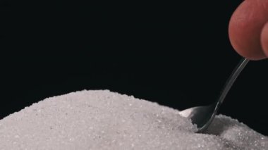 Bir çay kaşığı beyaz granüllü şekeri bir şeker yığınından ölç. Siyah arka planda bir kaşık dolusu şeker almak. Yakın plan. Bağımlılık, sağlıksız beslenme, aşırı kilo, diyabet, obezite
