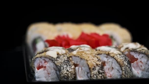 プラスチック製の箱に入った寿司は 黒い背景のクローズアップを回転させる デリバリーコンテナに異なる美味しい日本ロールのセット寿司ボックス 食糧配達 テイクアウト食品のコンセプト — ストック動画