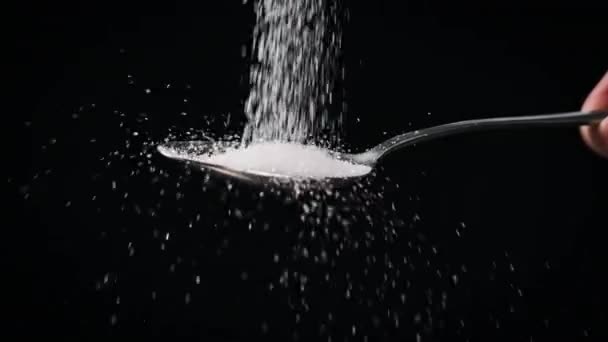 砂糖は黒い背景の遅い動きにスプーンに落ちる クローズアップ 白い粒状糖のテーブルスプーン1杯を測定する 砂糖中毒 不健康な食事 太りすぎ 糖尿病 肥満コピースペース — ストック動画